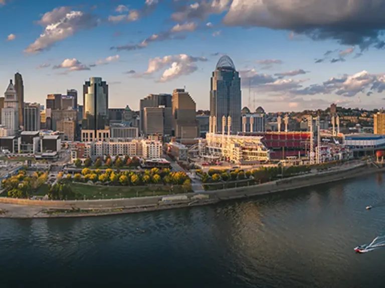 City skyline with a riverfront park and a large stadium under a partly cloudy sky.