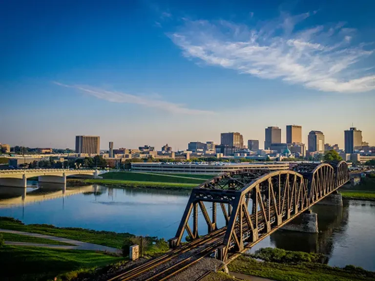 Steel truss bridge over a river with a city skyline under a blue sky.